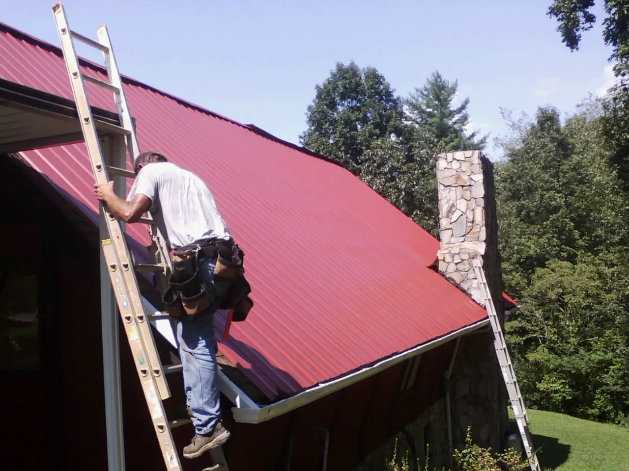 Roofer on a ladder beside a red metal roof and stone chimney — Soffit & Fascia Repair by Roofing Brothers in Millbrook